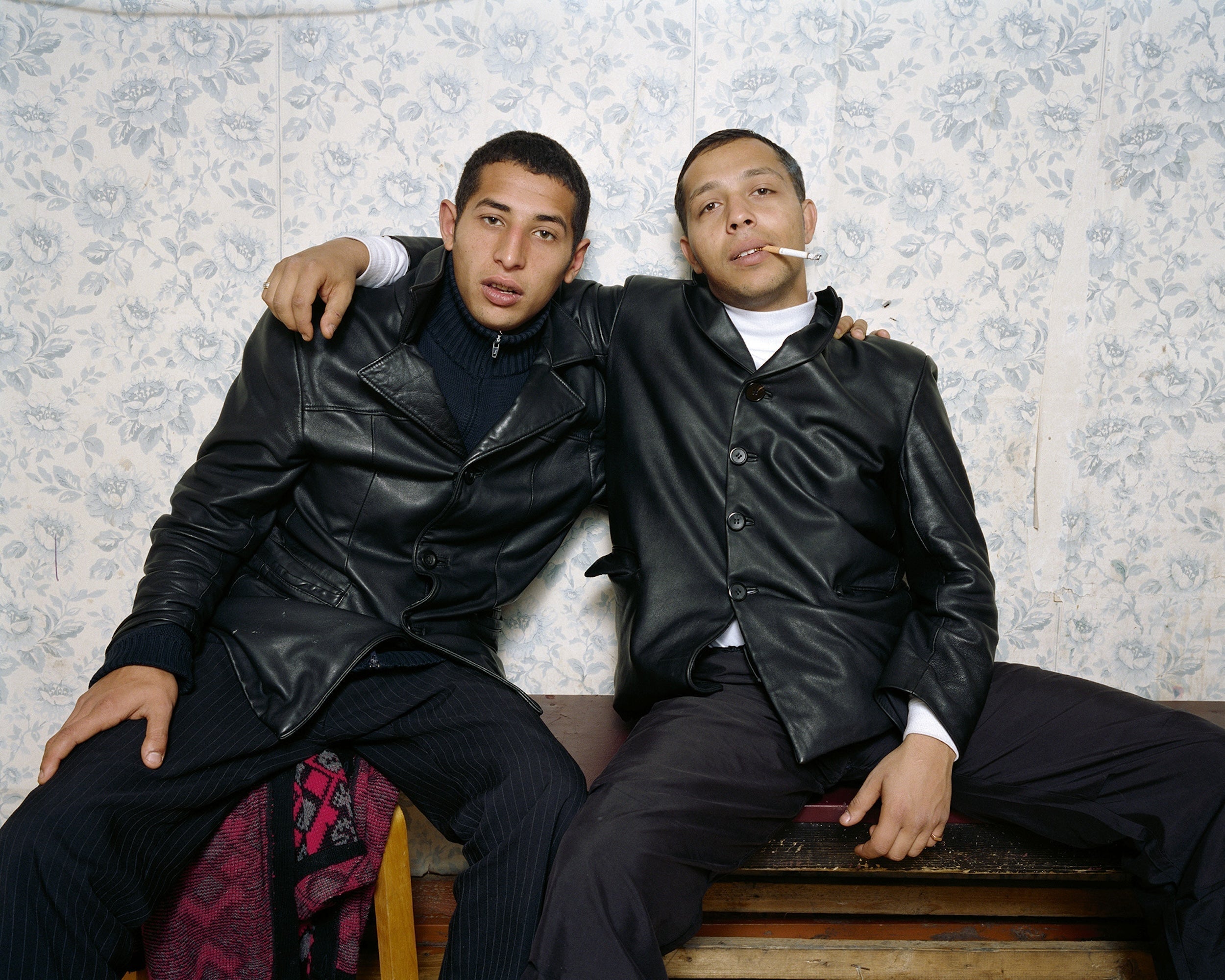 Two men wearing black leather jackets posing against floral wallpaper.