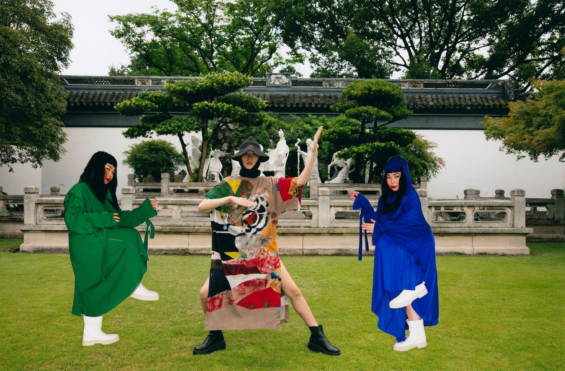 Three people in avant-garde costumes posing in front of a traditional white wall and garden.