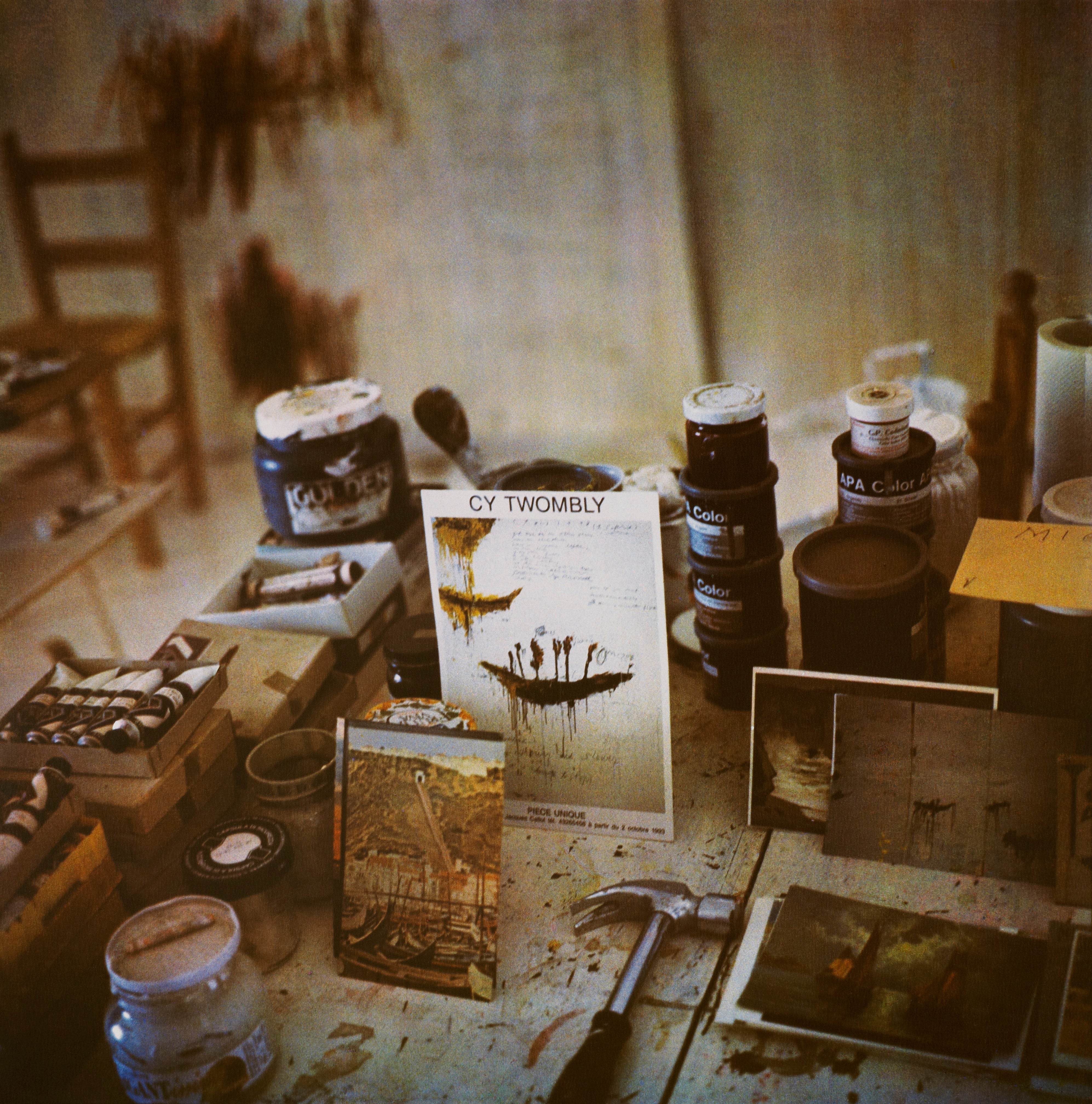 Artist's studio desk with paint jars, brushes, a hammer, and a Cy Twombly artwork.