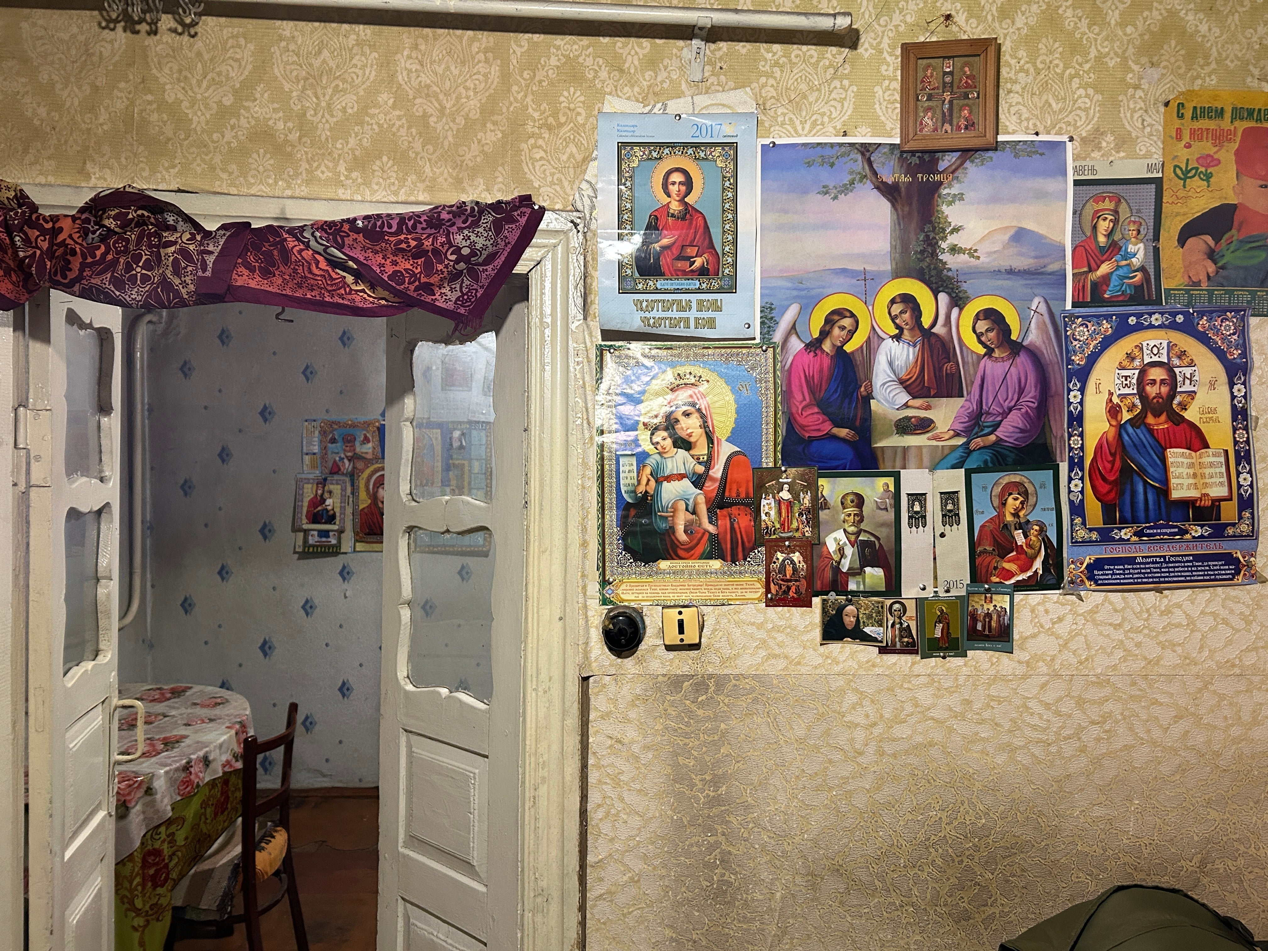 Various Orthodox Christian icons and posters displayed on a textured wallpaper wall next to a doorway.