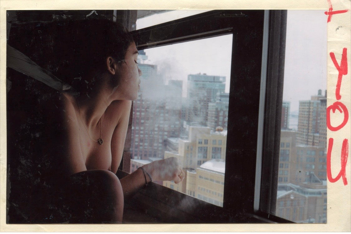 A woman looks out a window at a city skyline in a grainy, analog photograph.