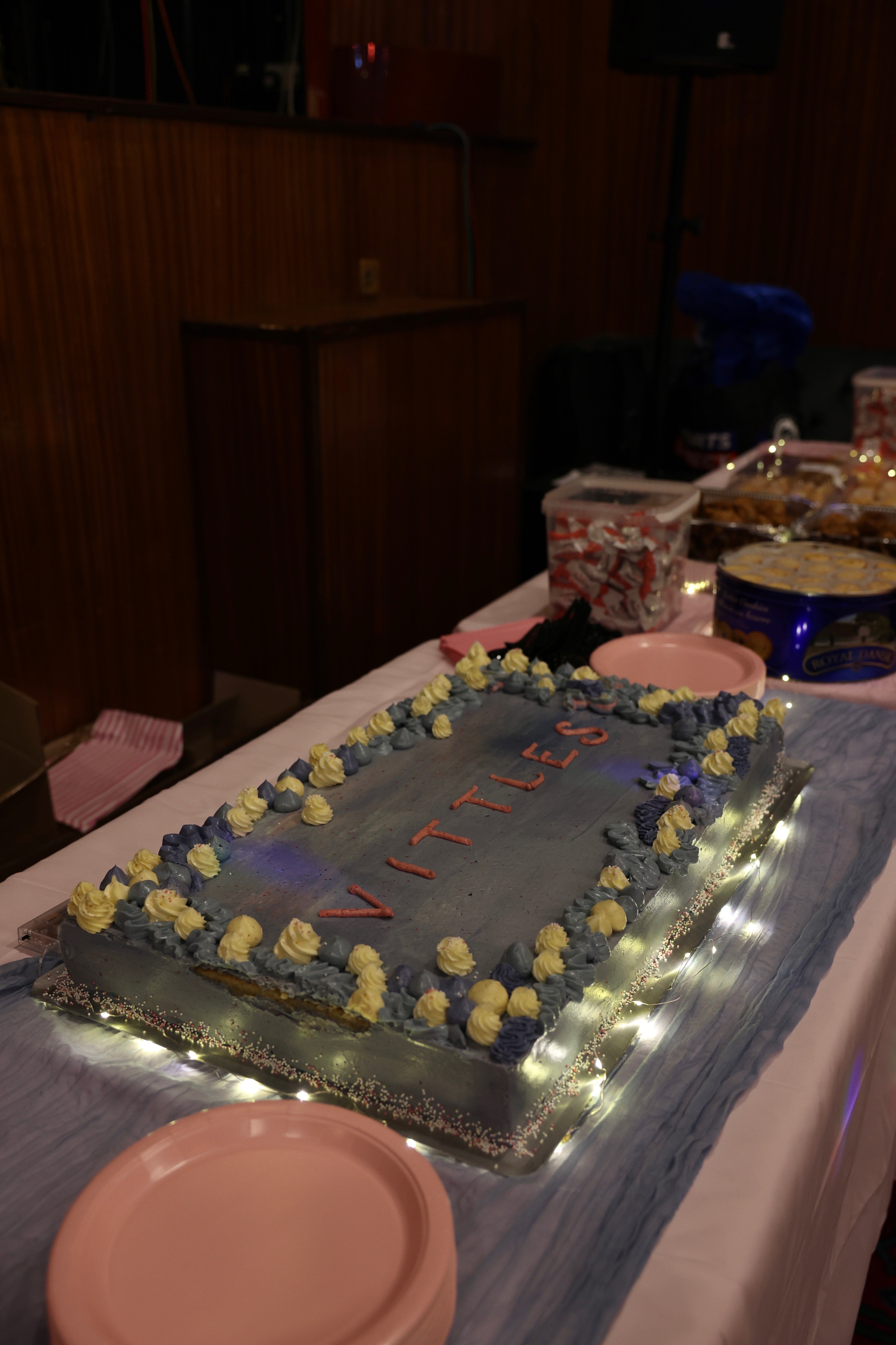 A rectangular blue frosted sheet cake with yellow and purple icing accents and the word VITLES.