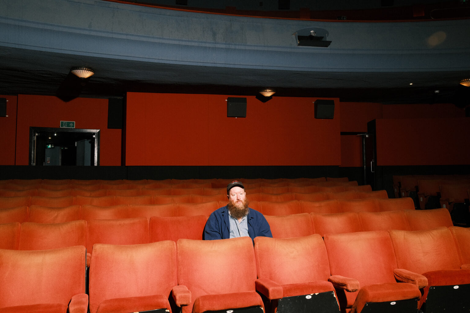 Bearded man sitting alone in rows of red velvet cinema seats.