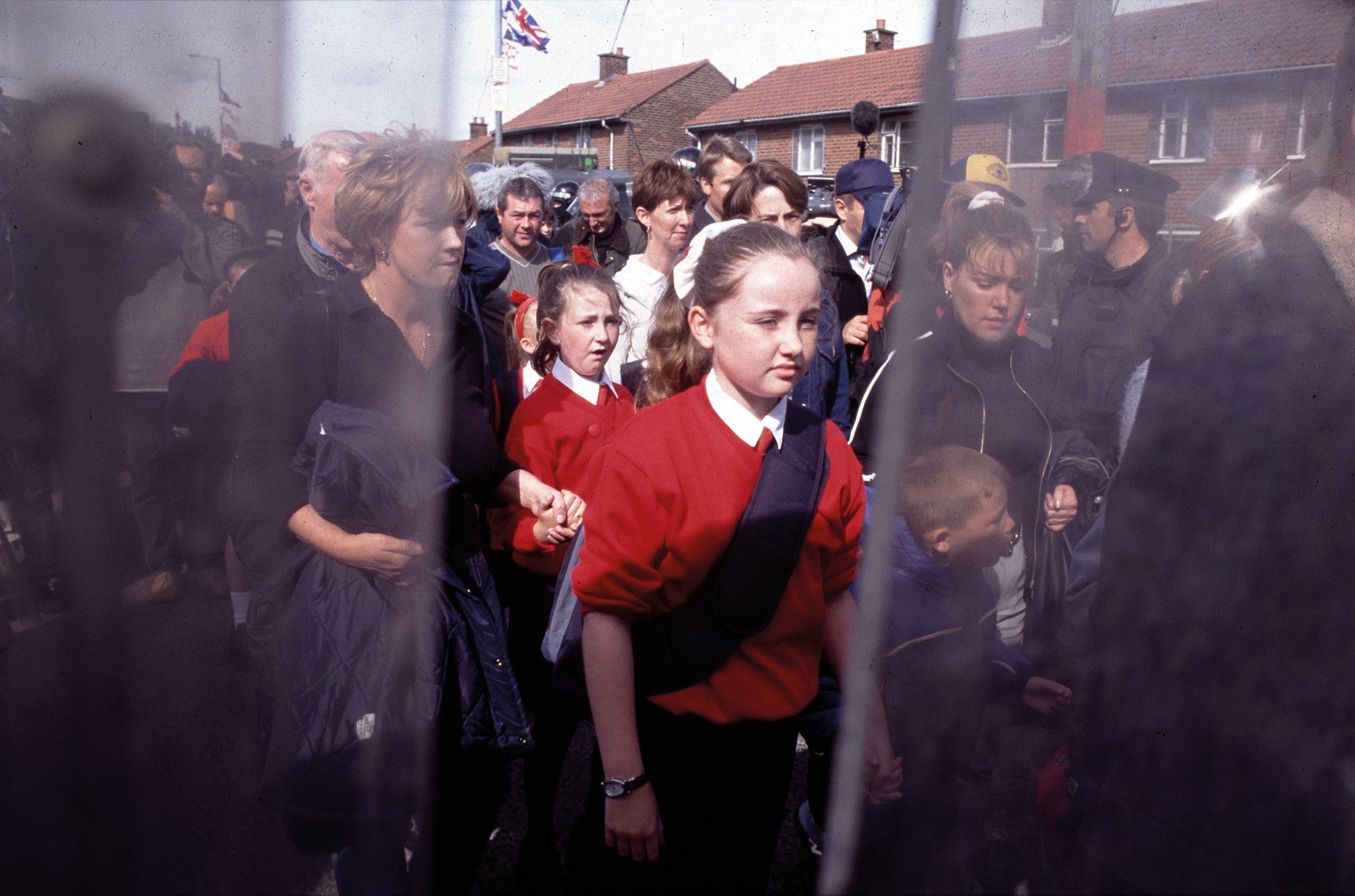 A group of people, including children in red uniforms, standing behind a fence outdoors.