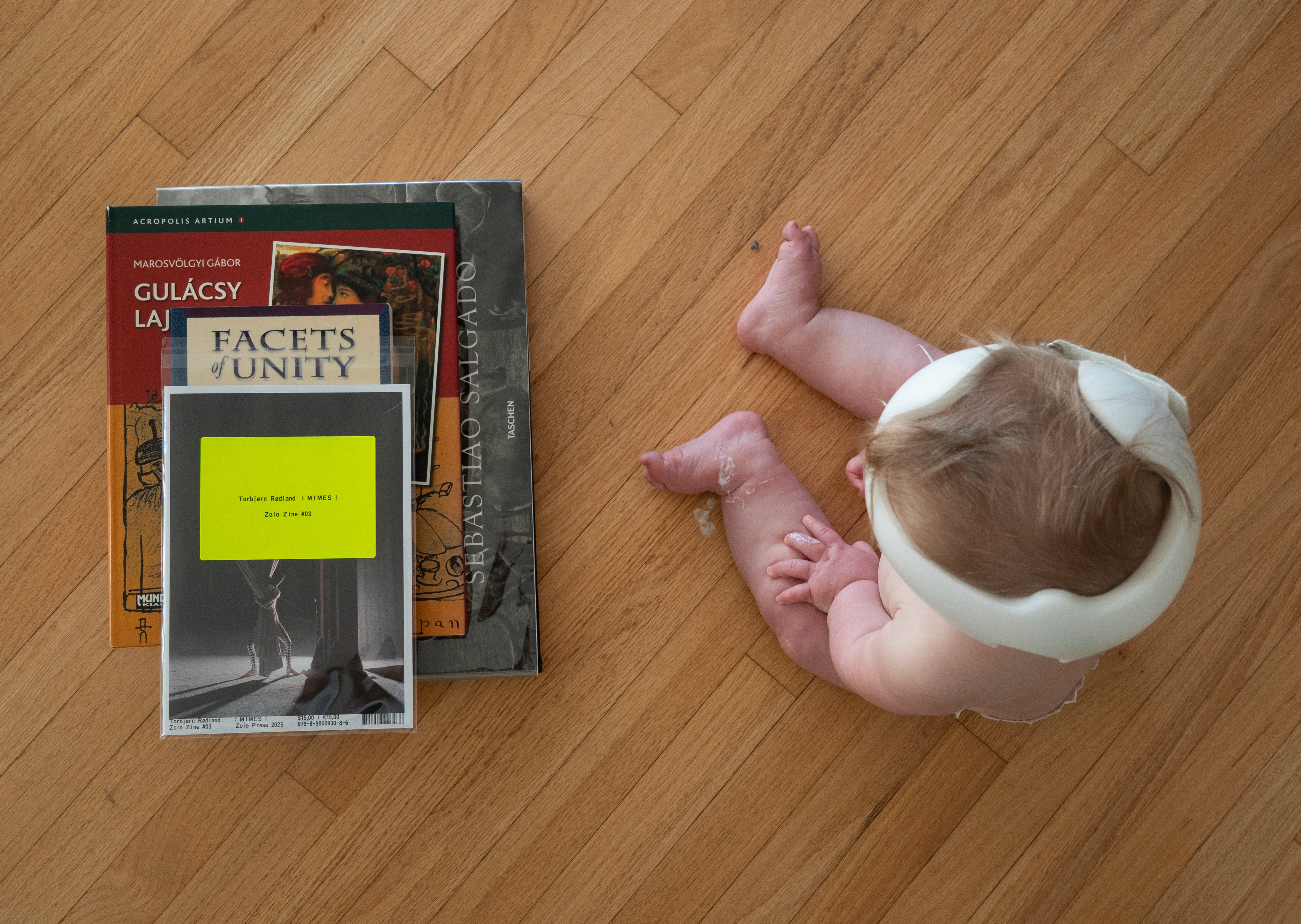 A baby wearing noise-canceling headphones lying on a wooden floor next to a stack of art books.