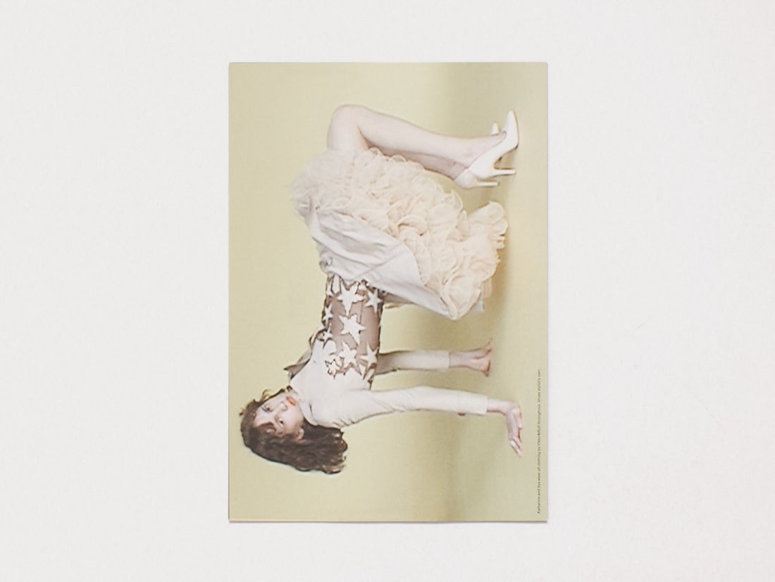 A woman posing horizontally in a white ruffled dress and star-patterned top against a pale yellow background.
