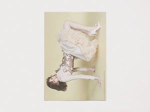 A woman posing horizontally in a white ruffled dress and star-patterned top against a pale yellow background.
